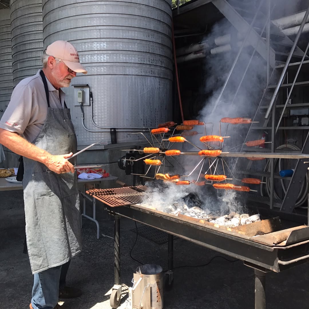 An older man in an apron grills multiple sausages on a unique vertical rack over a charcoal grill, smoke swirling into the air. Large metal tanks and a ladder in the background create an authentic atmosphere for The Cookout Collection—an experience best enjoyed with a chilled glass of Zinfandel Rosé.