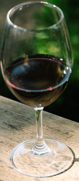 A glass of Hayne Vineyard - BTG red wine sits on a wooden surface outdoors, with green foliage blurred in the background.